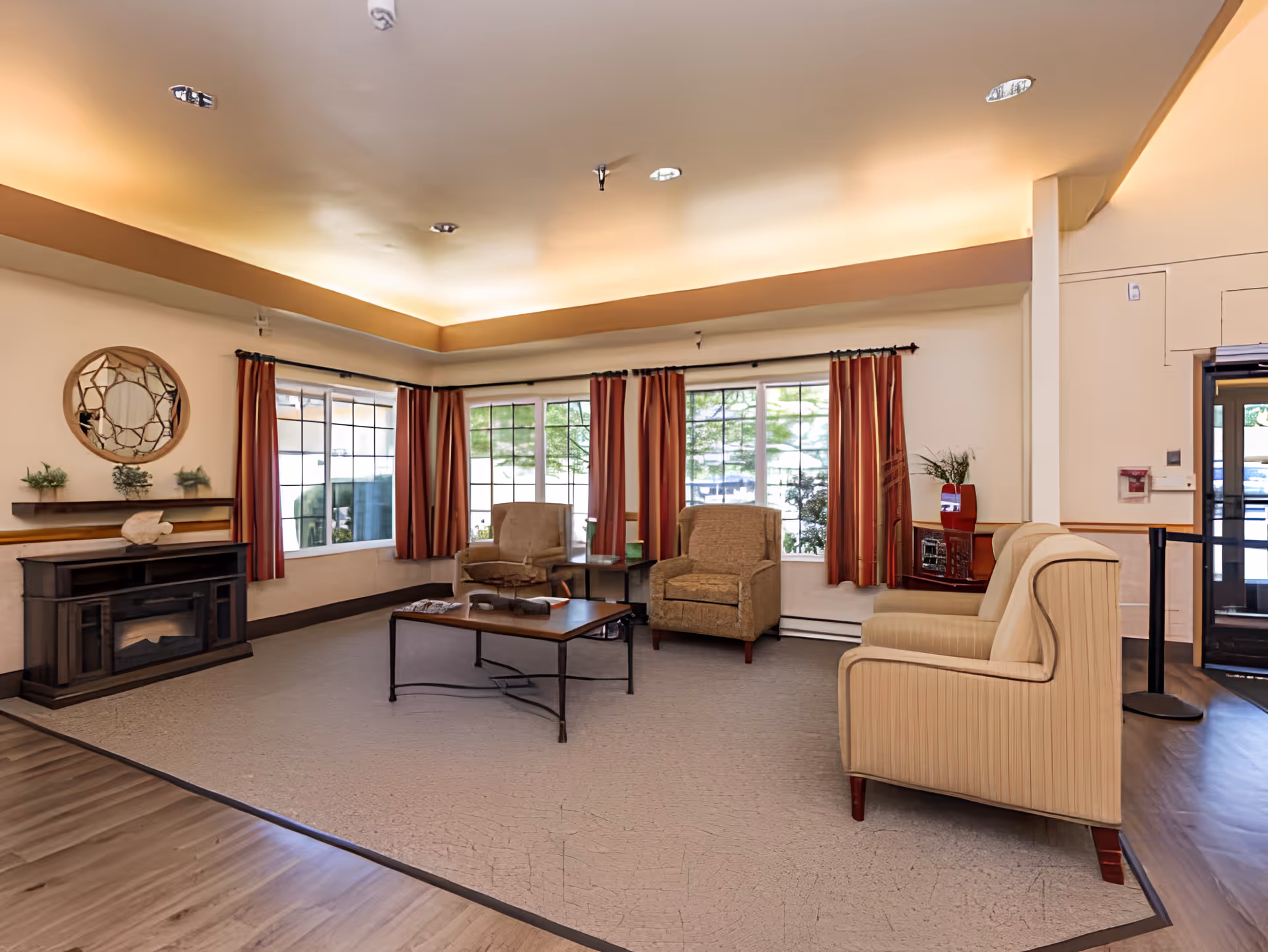 A cozy living room area with beige and brown upholstered chairs arranged around a wooden coffee table. The room has large windows with red curtains, a decorative round mirror on the wall, a small fireplace, and soft overhead lighting. There is a wooden floor partially covered by a large beige rug.