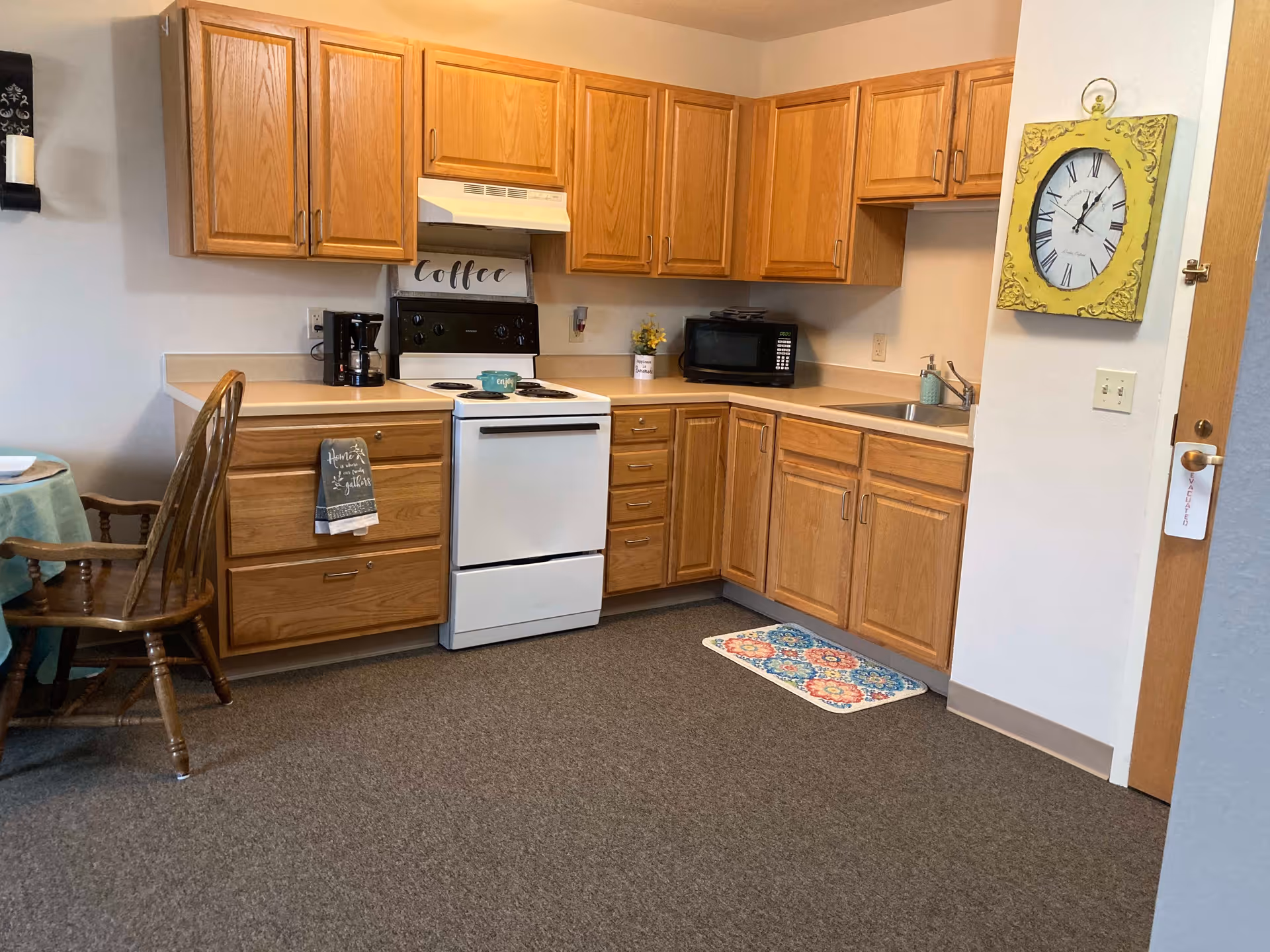 A kitchen area with wooden cabinets, a white stove with a coffee sign above it, a coffee maker, a microwave, a sink, and a colorful rug on the floor. There is a wooden chair and part of a dining table with a blue tablecloth visible on the left side. A yellow wall clock hangs on the right wall above a door with an evacuation tag on the handle.