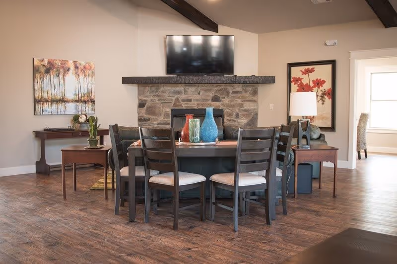 Communal living area with a round dining table and chairs in front of a stone fireplace topped by a mounted TV, decorated with vases and wall art.