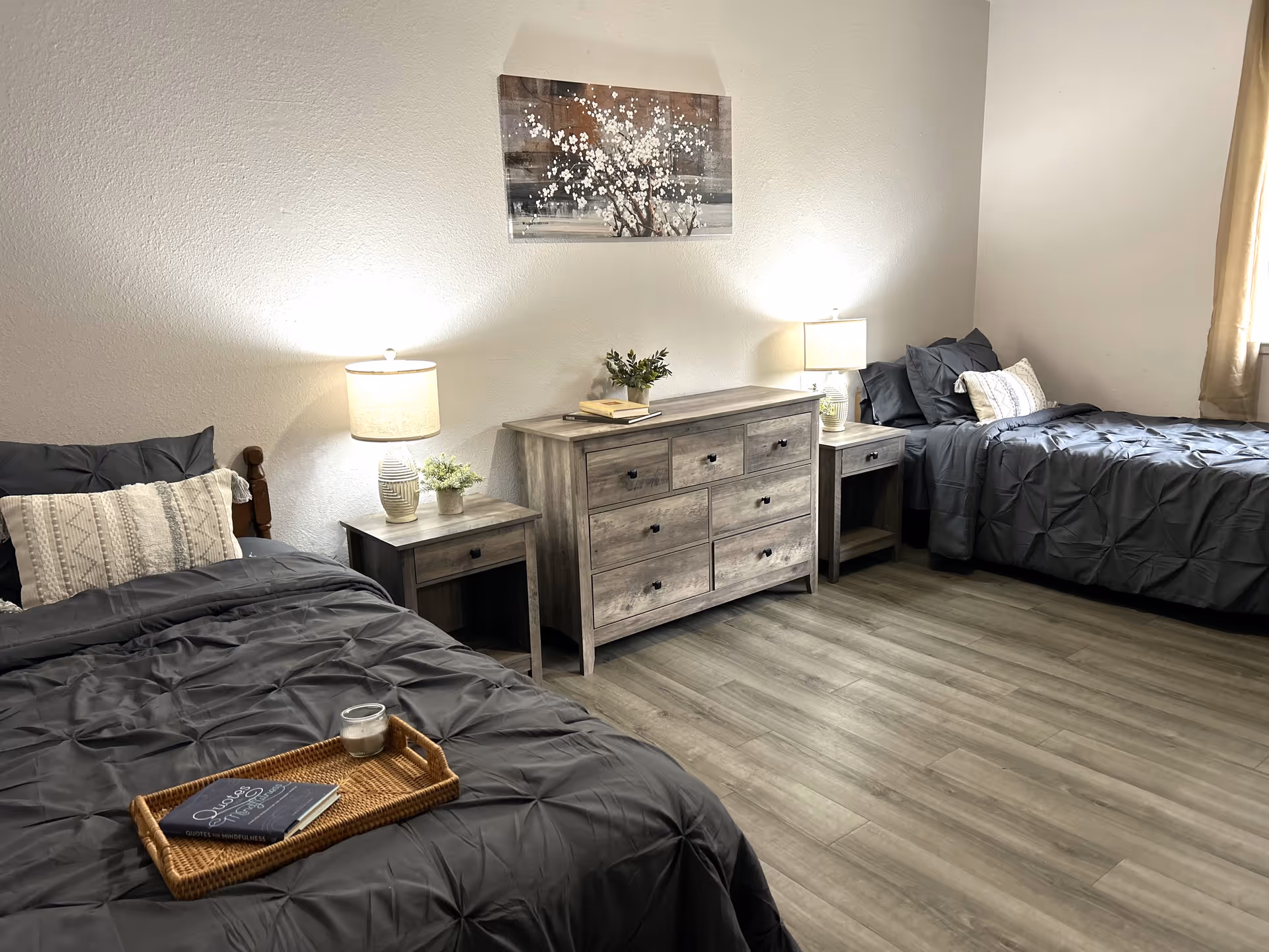 A bedroom with two twin beds, each with dark gray bedding and decorative pillows. Between the beds is a wooden dresser with six drawers, flanked by two matching nightstands, each with a lamp and a small plant. A framed artwork featuring white blossoms hangs on the wall above the dresser. The room has light-colored walls and wood flooring, with a window partially covered by beige curtains on the right side.
