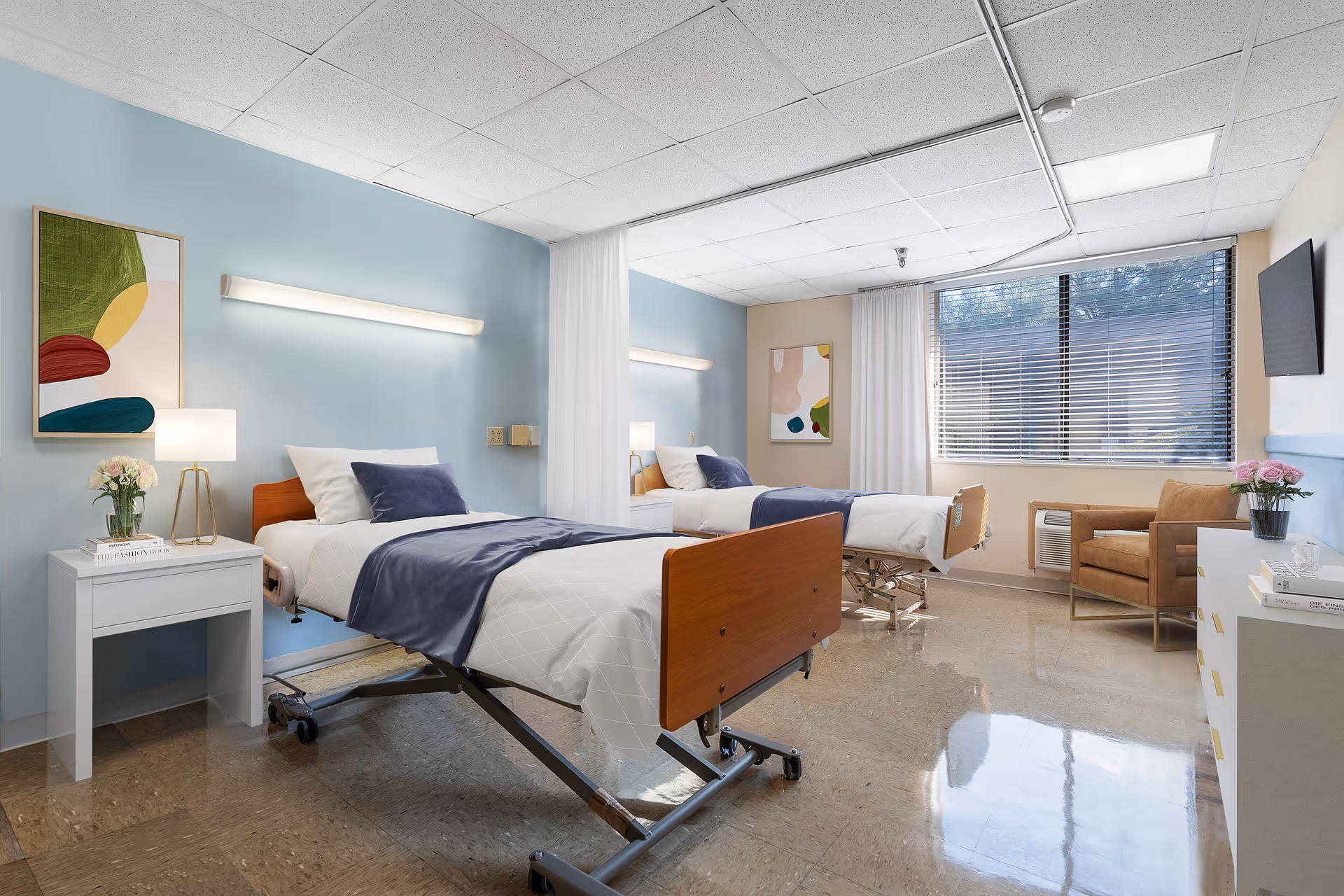 A bright shared nursing room with two hospital beds, bedside tables, chairs, and a large window.