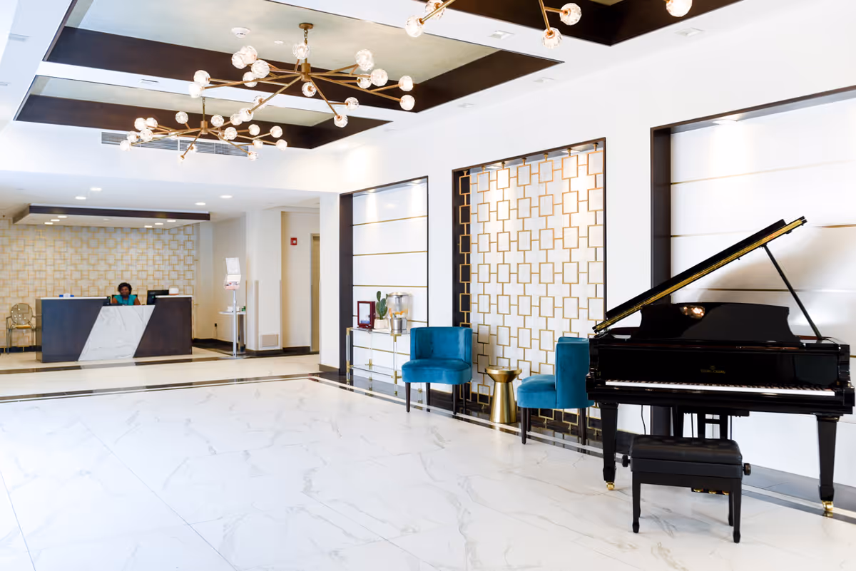 Bright and spacious lobby area with white marble flooring, a black grand piano with a matching bench on the right, two teal velvet chairs with a small gold side table between them, and a reception desk with a person seated behind it in the background. The walls feature decorative gold geometric patterns and modern light fixtures hang from the ceiling.