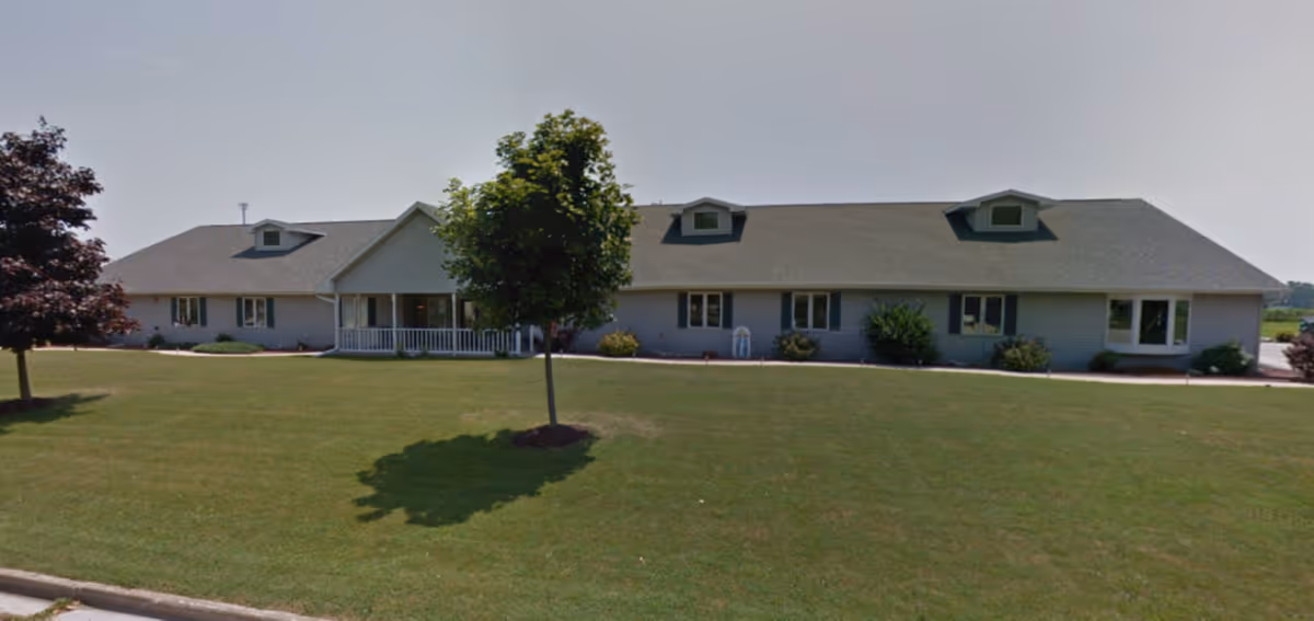 Single-story ranch-style senior living building with a front porch, dormer windows, and a large grassy lawn with a few trees.