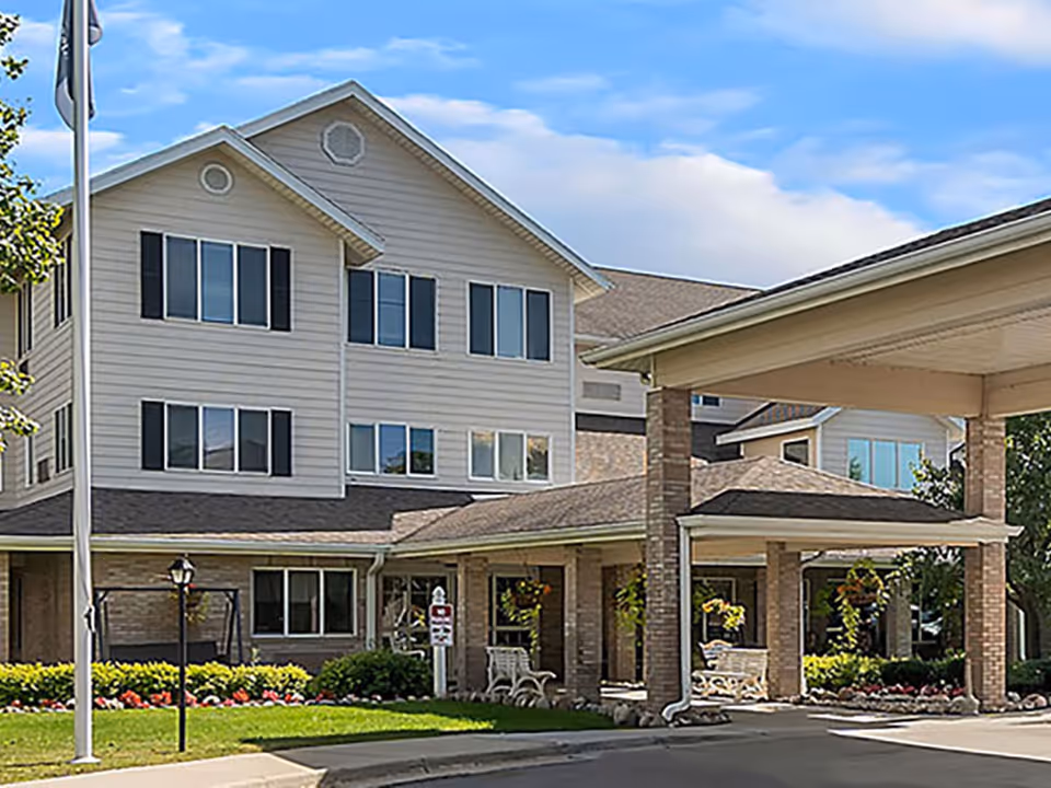 Exterior view of a multi-story senior living facility building with beige siding and black window shutters. The entrance features a covered drop-off area supported by brick columns, with benches and hanging flower baskets. The surrounding area has well-maintained landscaping with green grass, bushes, and flowers under a partly cloudy blue sky.