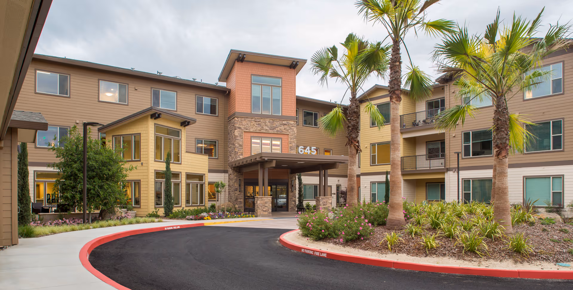 Front exterior view of Prairie City Landing facility showing a three-story building with a covered entrance, multiple windows, palm trees, and landscaped plants along a curved driveway.