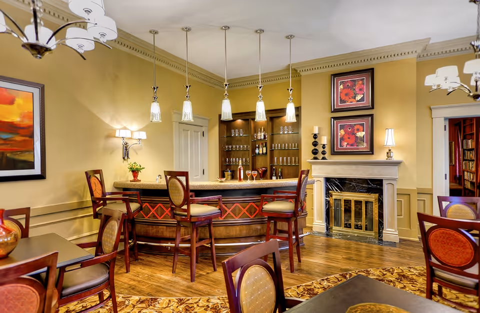 A cozy and elegant interior room featuring a wooden bar with five high chairs, pendant lights hanging above the bar, and shelves stocked with glasses and bottles. The room has warm yellow walls, a fireplace with a marble surround and brass screen, two framed floral artworks above the fireplace, and several tables and chairs with cushioned seats. The floor is wooden with a patterned rug partially visible.