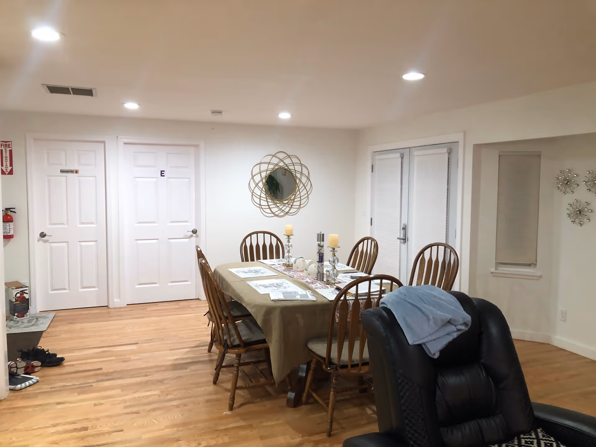 Interior dining area with a table set for meals surrounded by wooden chairs, a decorative wall mirror, and a black recliner in the foreground.