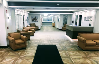 A spacious and well-lit reception area of Birchwood Terrace Rehab and Healthcare featuring comfortable brown armchairs and sofas arranged along the walls. The floor has a patterned design, and the reception desk is on the right side with the facility's name and logo displayed on the wall behind it. The area has a clean, modern look with white walls and ceiling lights.