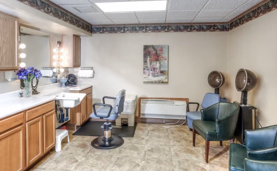 Interior view of a salon area in a senior living facility with a styling chair in front of a counter with a sink, mirror, and light bulbs. There are two hair drying chairs with hooded dryers on the right side, additional seating, a floral painting on the wall, and a heating/cooling unit below the painting.