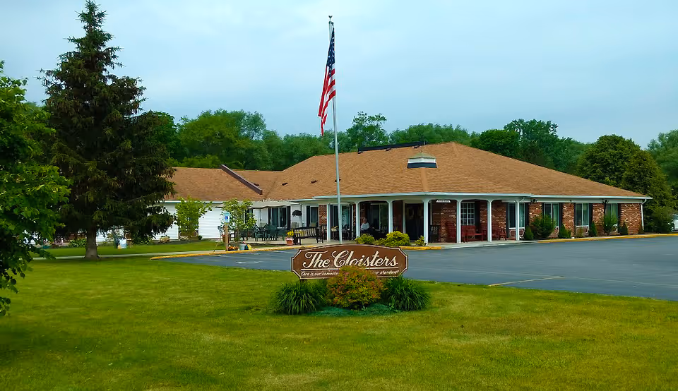 Exterior view of The Cloisters senior living facility showing a single-story brick building with a brown roof, a flagpole with an American flag, a parking lot, and a green lawn with trees and shrubs.