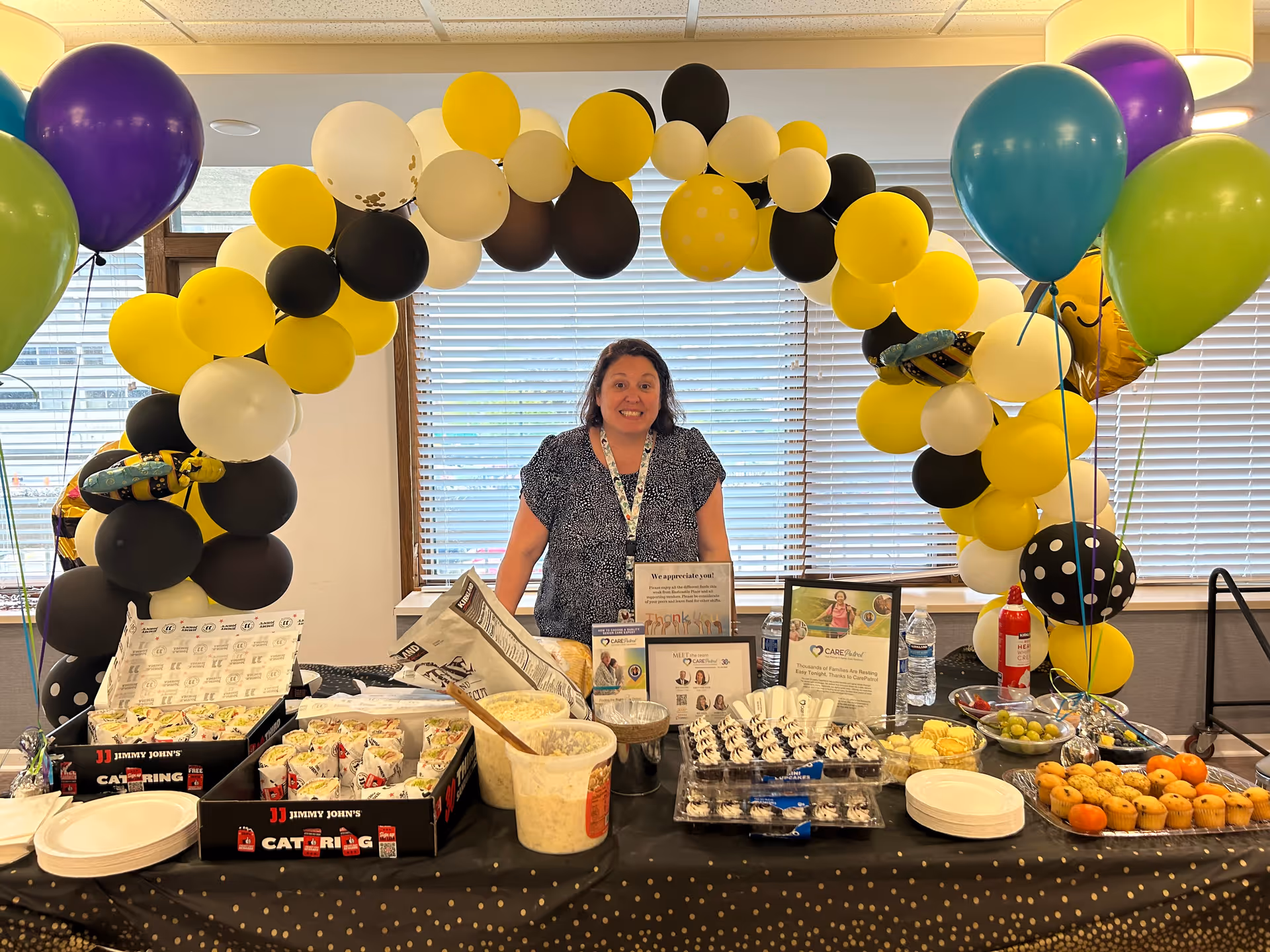 A woman stands behind a table filled with various food items including sandwiches, cupcakes, muffins, grapes, and other snacks. The table is decorated with a black tablecloth with gold dots. Behind her is an arch made of yellow, black, white, and a few polka-dotted balloons. Additional balloons in purple, green, and blue are on either side of the table. The setting appears to be indoors with large windows and blinds in the background.