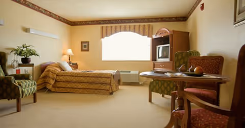 A cozy bedroom in a senior living facility with a single bed covered in a patterned bedspread, a nightstand with a lamp and a plant, two upholstered chairs, a wooden table with fruit and a magazine, a TV cabinet with a small television, and a large window with a valance letting in natural light.