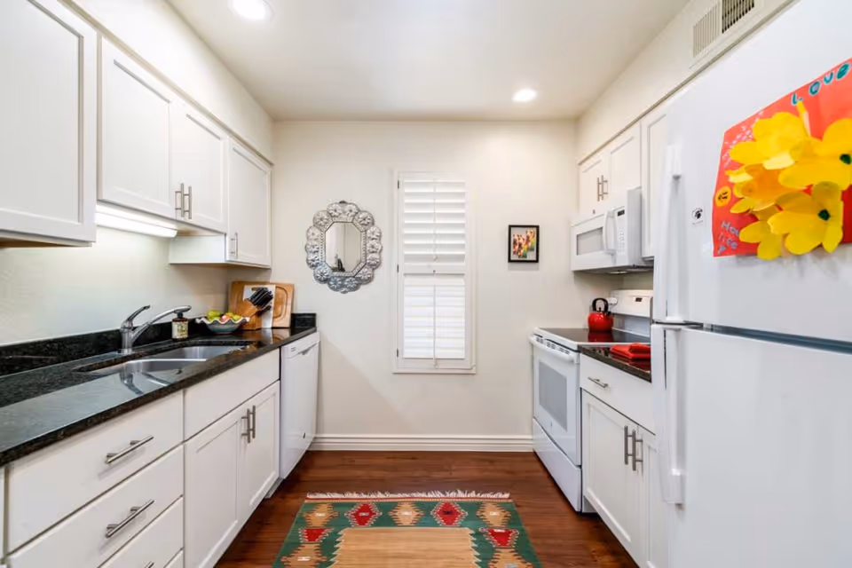 Bright galley kitchen with white cabinets, black countertops, white appliances, and a colorful rug.
