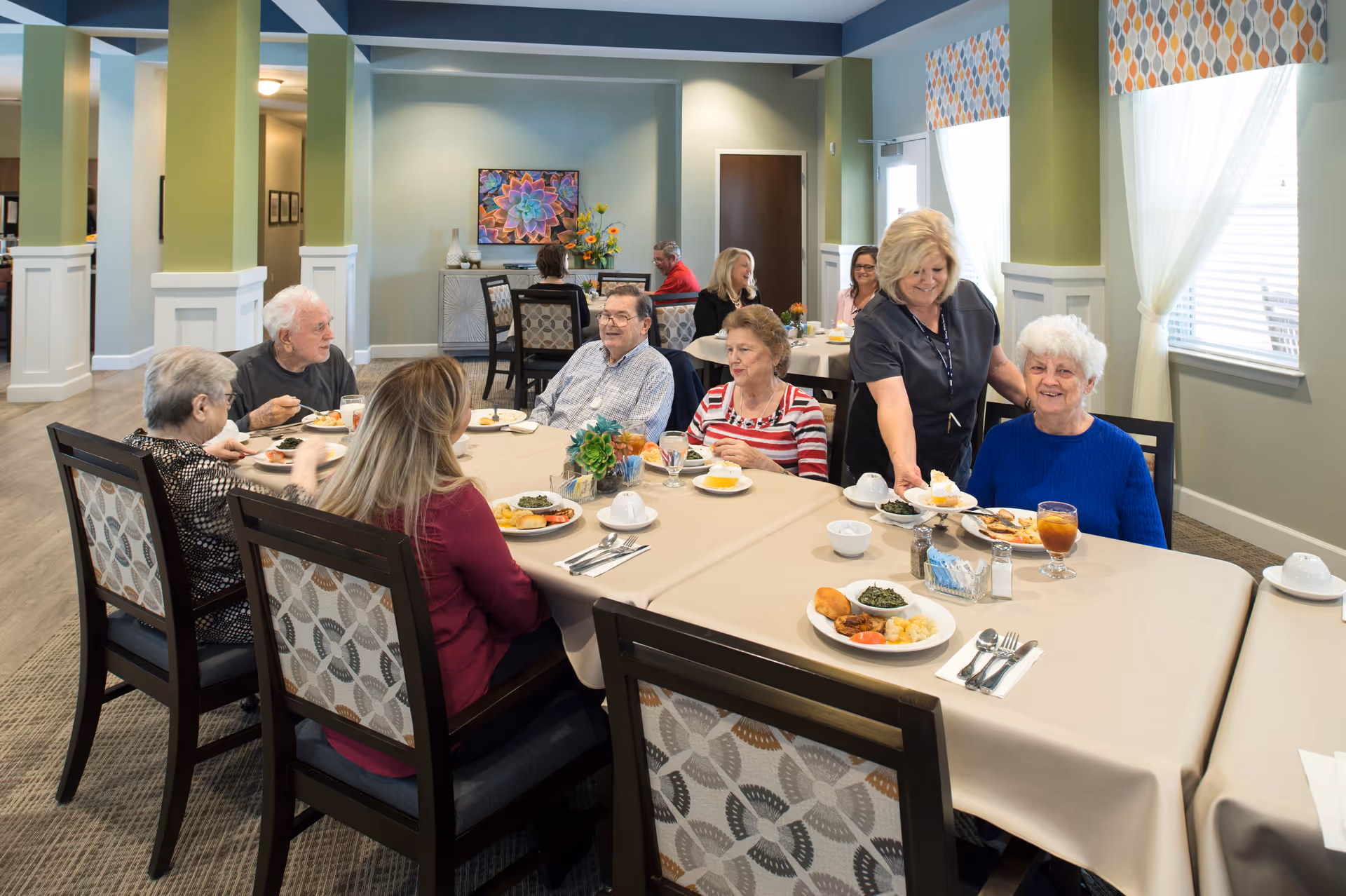 A group of elderly people seated around a dining table in a well-lit room with green walls and large windows. A caregiver is serving food to one of the elderly women wearing a blue sweater. Plates of food and drinks are on the table, and other residents are seen eating and conversing in the background.