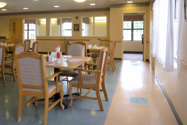 A dining area with wooden tables and cushioned chairs arranged neatly. The tables have placemats, condiments, and napkin holders. The room has large windows with curtains allowing natural light to enter, and a hallway is visible in the background.