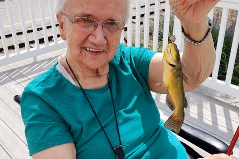 An elderly woman wearing glasses and a teal shirt is sitting outdoors on a wooden deck, smiling and holding up a small yellow fish she caught. There is a white railing behind her and some greenery in the background.