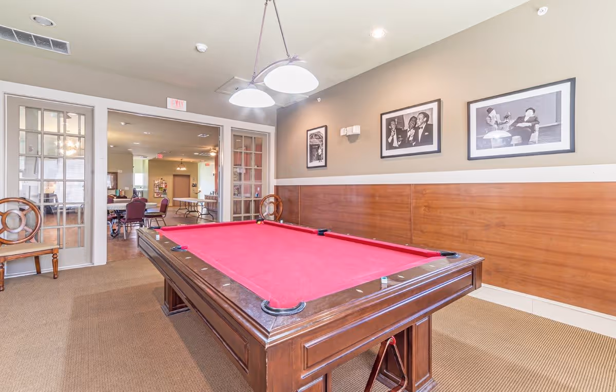 Interior room with a red felt pool table in the center, wooden chairs along the walls, and framed black and white photos hanging on the beige wall. Through glass double doors, another room with tables and chairs is visible.