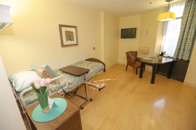 A bright room with a single hospital-style bed covered with patterned bedding and a pillow. Next to the bed is a small wooden nightstand with a vase of pink tulips on a turquoise placemat. A brown leather chair and a dark wooden desk with a small plant and a plate are positioned near a window with light blue curtains. A wall-mounted TV and a clock are visible on the far wall. The floor is light wood, and the walls are painted pale yellow.