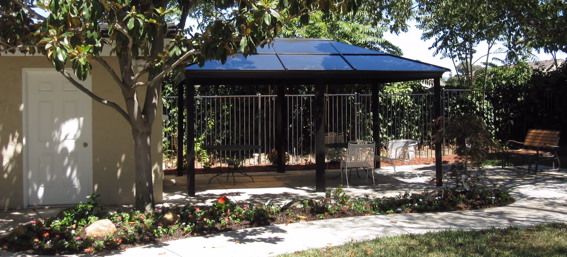 Outdoor patio area with a black metal gazebo featuring a solar panel roof, surrounded by trees and greenery. There are metal chairs and tables under the gazebo, a wooden bench to the right, and a flower bed with colorful flowers in the foreground. A beige building with a white door is visible on the left side.