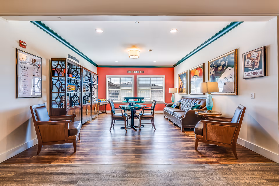 A bright and spacious game room in a senior living facility with wooden flooring, a brown leather sofa, two wooden armchairs, a round table with four chairs, and a large window letting in natural light. The walls are decorated with framed artwork and signs indicating it is a game room.