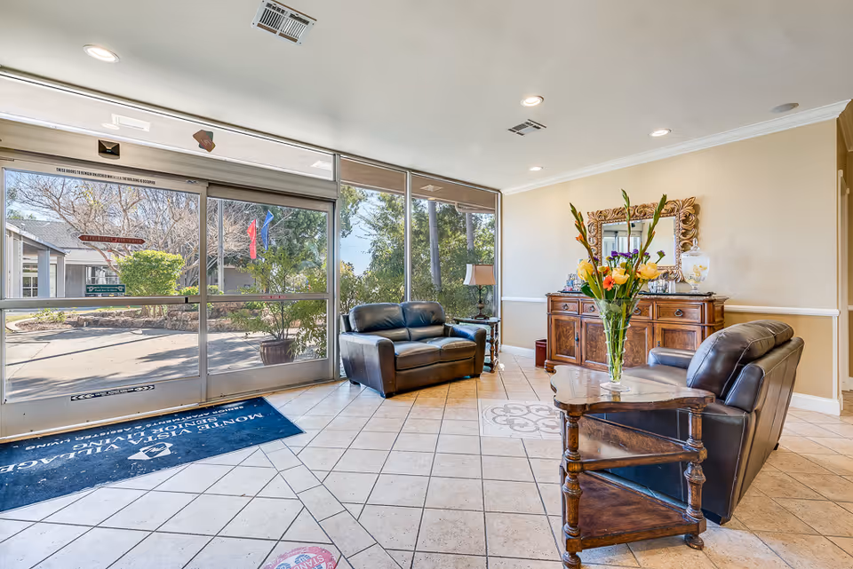 A bright and welcoming lobby area of Monte Vista Village featuring large glass sliding doors that open to an outdoor view with trees and flags. Inside, there are two dark leather armchairs, a wooden side table with a vase of colorful flowers, a wooden cabinet with a decorative mirror above it, and a floor mat with the facility's name.