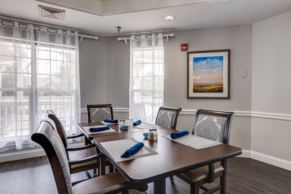 A dining room with a rectangular wooden table set for five people. Each place setting has a beige placemat and a rolled blue napkin. The chairs have patterned upholstery. The room has light gray walls, two large windows with sheer white curtains, a framed landscape painting on the wall, and a fire alarm above it.