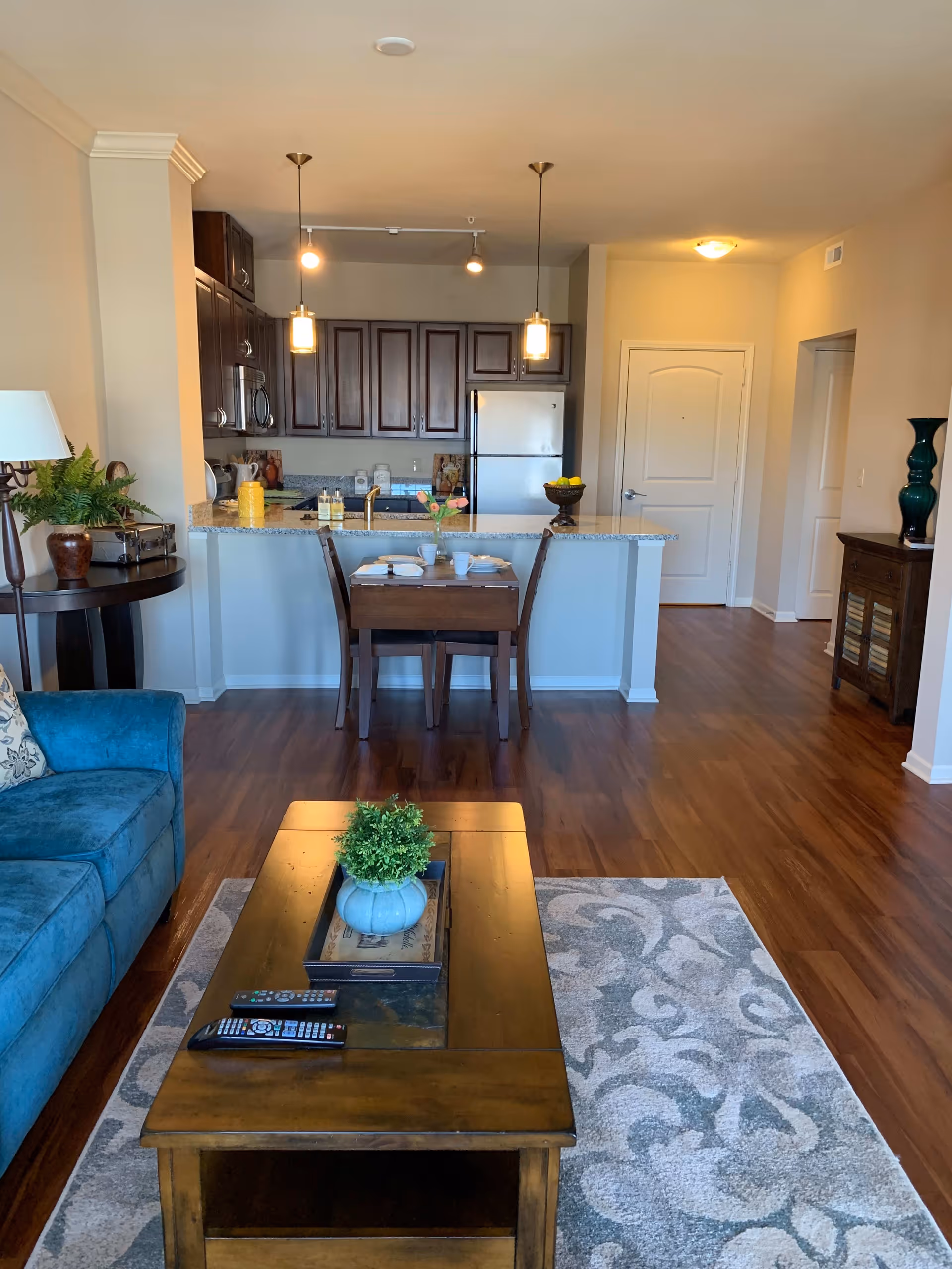 View of a cozy living room and kitchen area in an apartment at McCrite Plaza at Briarcliff. The living room features a blue sofa, a wooden coffee table with a small plant and remote controls on it, and a patterned area rug. The kitchen has dark wood cabinets, a granite countertop with a small dining table set for two, pendant lights, and stainless steel appliances including a refrigerator and microwave. The flooring is wood, and there are decorative items like a lamp, plants, and vases around the space.