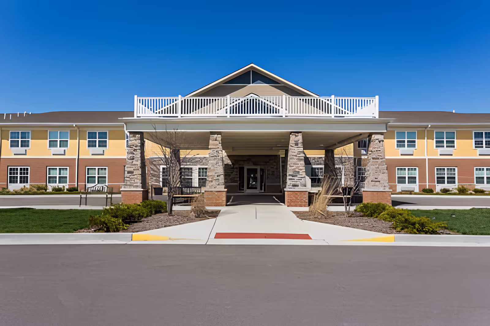 Front exterior view of Traditions at Reagan Park building with a covered entrance supported by stone pillars, a paved driveway, and a clear blue sky.