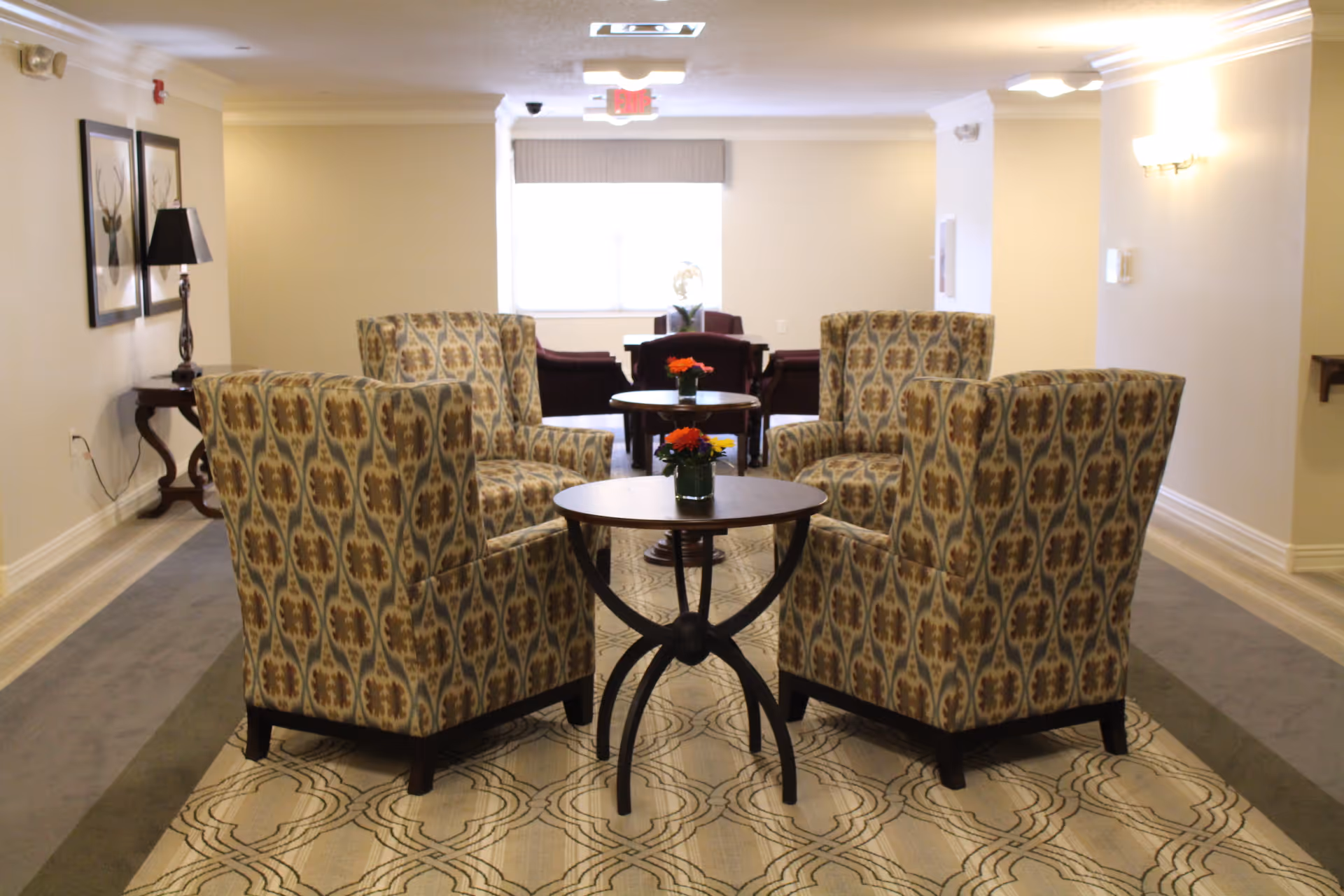 A seating area in an assisted living facility with four patterned armchairs arranged around a small round wooden table with a small flower arrangement. In the background, there are additional chairs and tables near a window with blinds. The walls are light-colored, and the floor has a patterned carpet.