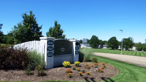 Outdoor view of the entrance sign for Stow Glen Retirement Village surrounded by landscaped plants and flowers, with a clear blue sky and green trees in the background.