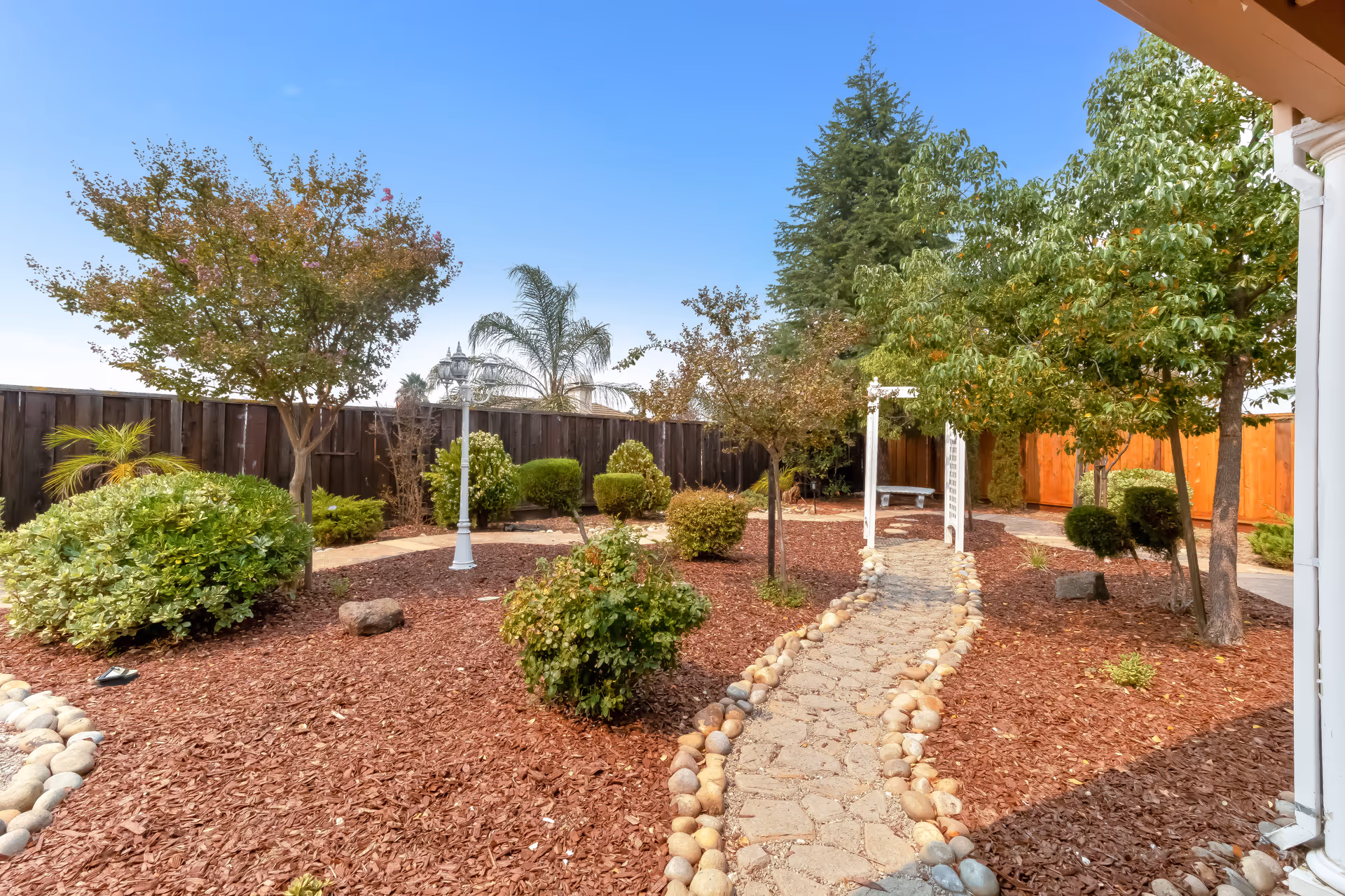 A landscaped outdoor garden area with a stone pathway bordered by round stones winding through mulch-covered ground. The garden features various green shrubs and trees, a white lamppost, and a white wooden pergola with a bench underneath. A wooden fence encloses the garden under a clear blue sky.