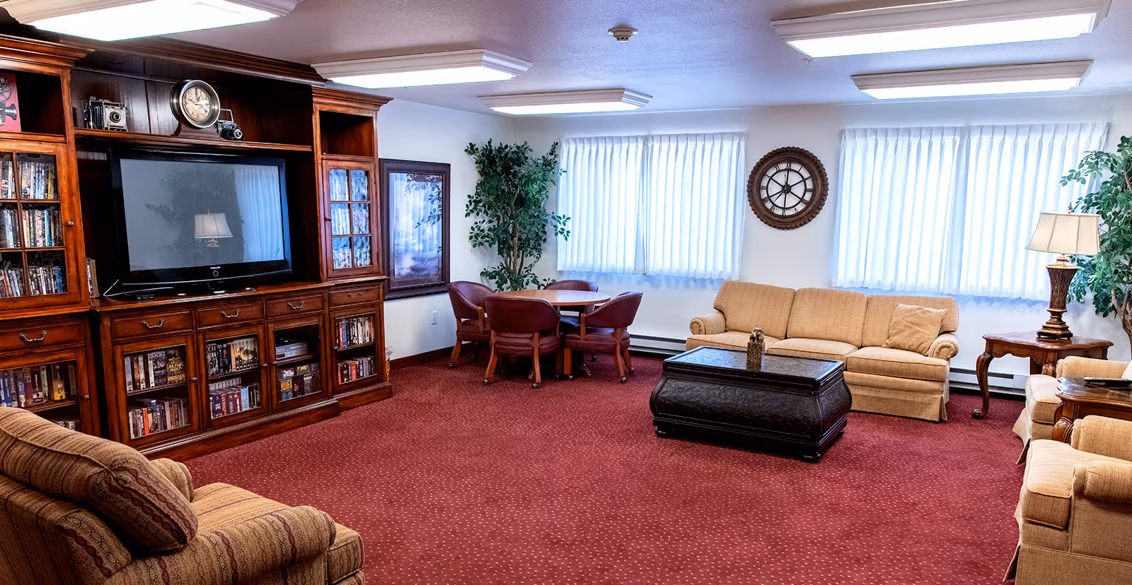 Cozy common room with sofas and armchairs, a large wooden entertainment cabinet with a TV and books, a coffee table, and a small table with chairs.