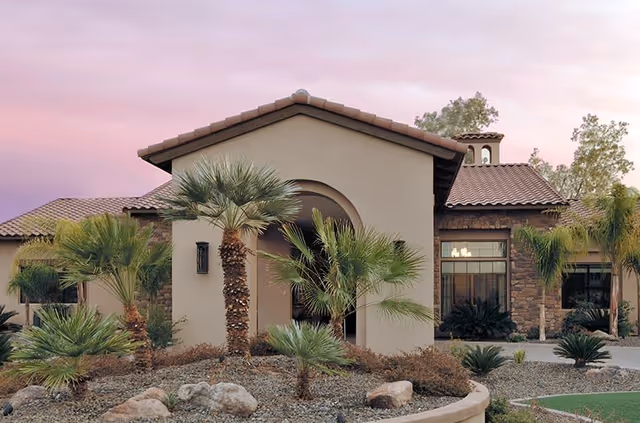 Exterior view of a single-story building with a tiled roof and stone accents, surrounded by desert landscaping including palm trees and rocks, under a pink and purple sky at sunset.