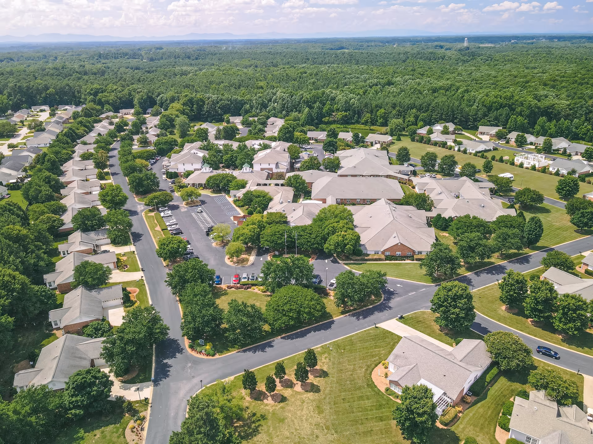 Aerial view of Summit Hills senior living facility showing multiple buildings with gray roofs surrounded by green trees and lawns, parking lots with cars, and roads connecting the buildings. The background features a vast forested area under a partly cloudy sky.