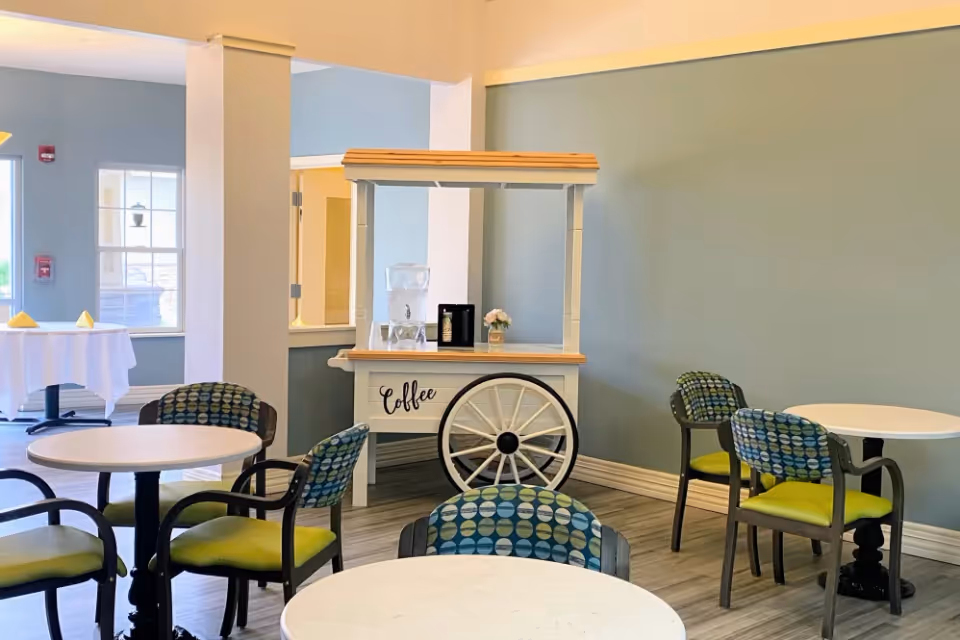 Dining room with round tables, green-and-blue patterned chairs, and a small coffee cart with a decorative wheel against a pale blue wall.