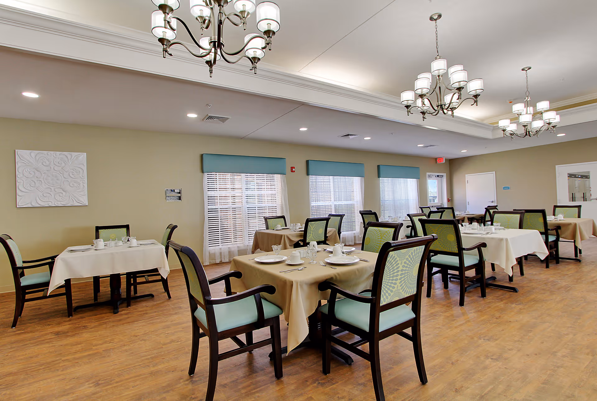 Bright dining room with multiple tables set with plates and cups, upholstered chairs, chandeliers, and large windows with blinds.