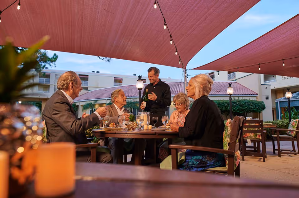 A group of elderly people sitting around a round outdoor dining table under a canopy with string lights. A waiter is standing and pouring wine for them. The setting is in the courtyard of a senior living facility with buildings and greenery in the background.