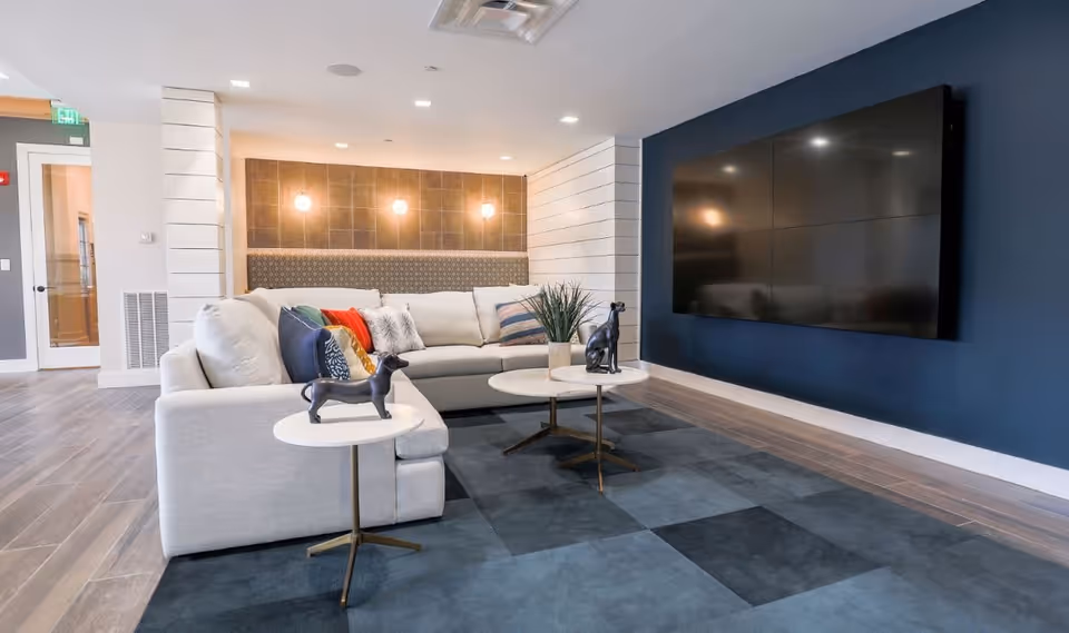 Modern living room with a large white sectional sofa adorned with colorful pillows, two small round white tables with dog sculptures, a large flat-screen TV mounted on a dark blue wall, and a patterned rug on the floor. The background features a wall with three round lights and a door leading to another room.