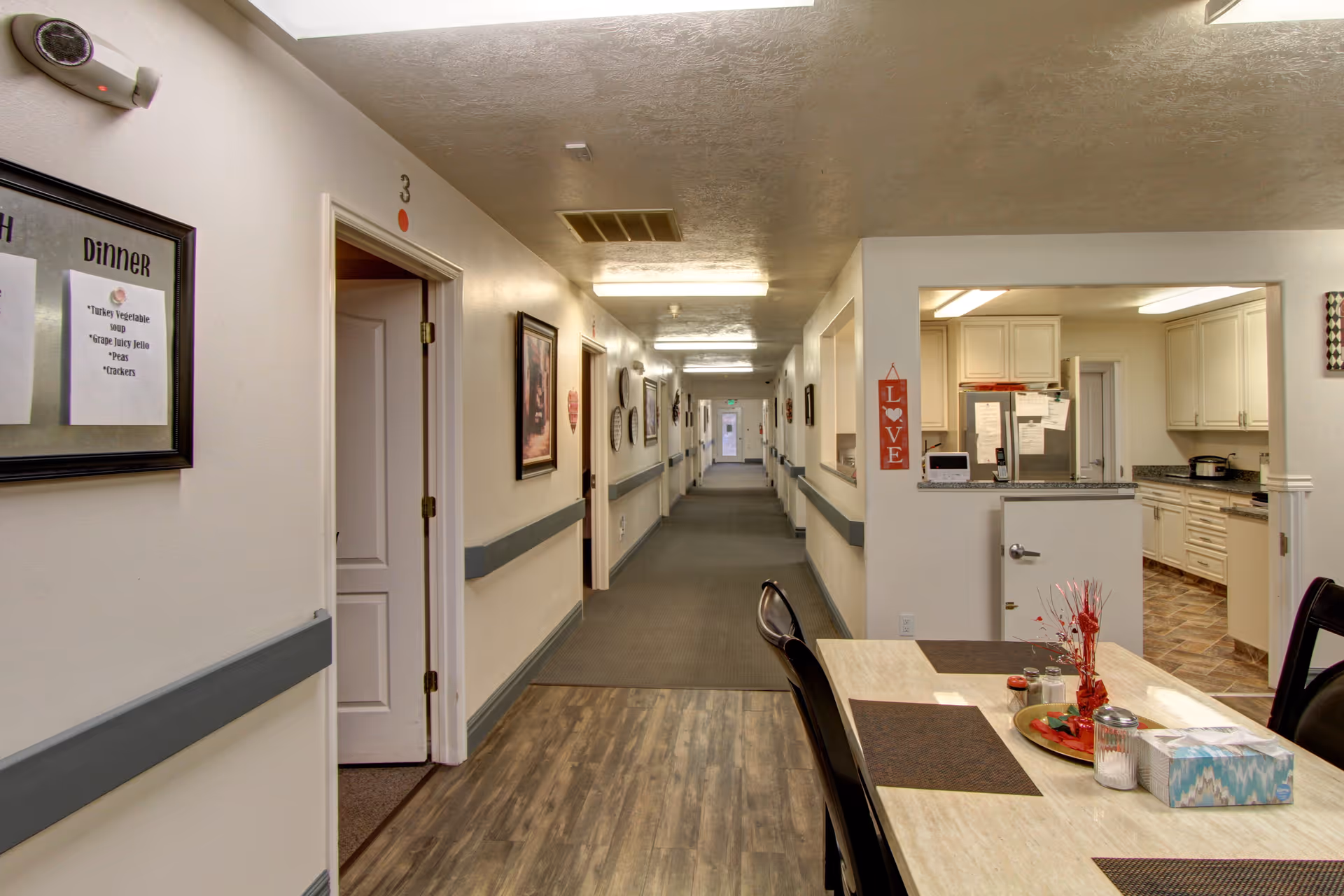Long interior hallway of a senior living facility with a dining table and a kitchen area visible on the right.