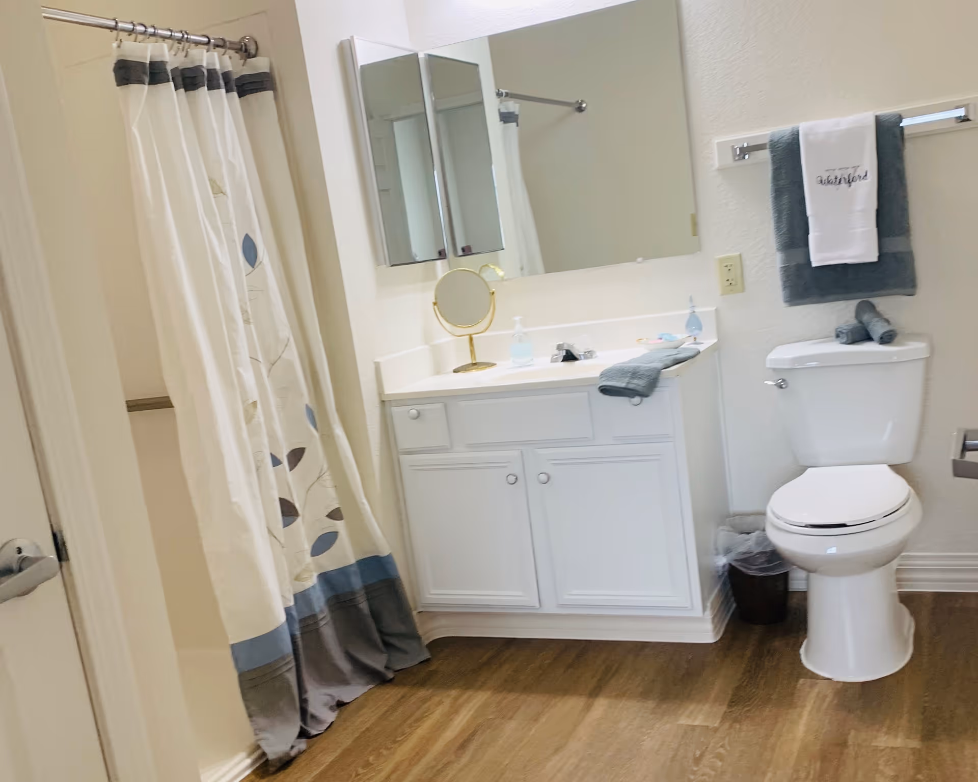 A clean bathroom featuring a white vanity with a large mirror above it, a small round makeup mirror, and a soap dispenser on the countertop. To the right is a white toilet with folded towels on top and a towel rack above holding a dark gray towel and a white towel embroidered with the word 'Waterford'. On the left side, there is a shower with a curtain decorated with a leaf pattern in shades of blue and gray. The floor is wood-style laminate.