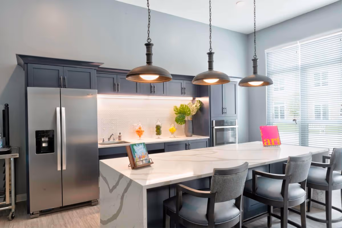 Modern kitchen with dark blue cabinets, a large white marble island with three gray cushioned chairs, three pendant lights hanging above the island, a stainless steel refrigerator, built-in oven, and a window with blinds letting in natural light.
