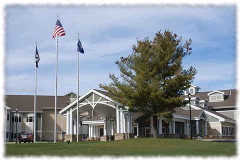 Exterior view of the New York State Veterans Home building with a covered entrance, three flagpoles displaying the American flag and other flags, a large tree, and a well-maintained lawn under a partly cloudy sky.