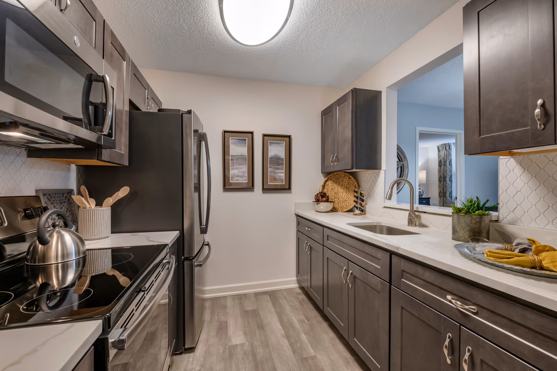 Modern kitchen with dark wood cabinets, stainless steel appliances including a microwave, stove, and refrigerator. The kitchen has a white countertop with a sink and faucet, decorative items such as a plant, a bowl, and a tray with napkins. Two framed pictures hang on the wall at the end of the kitchen. The floor is light wood, and there is a pass-through window to an adjacent room.