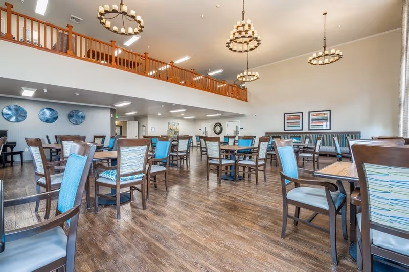Spacious dining room with wooden floors, multiple tables and chairs with blue and patterned cushions, high ceilings with hanging chandeliers, and a balcony railing above. The walls are decorated with framed artwork and circular wall decor.