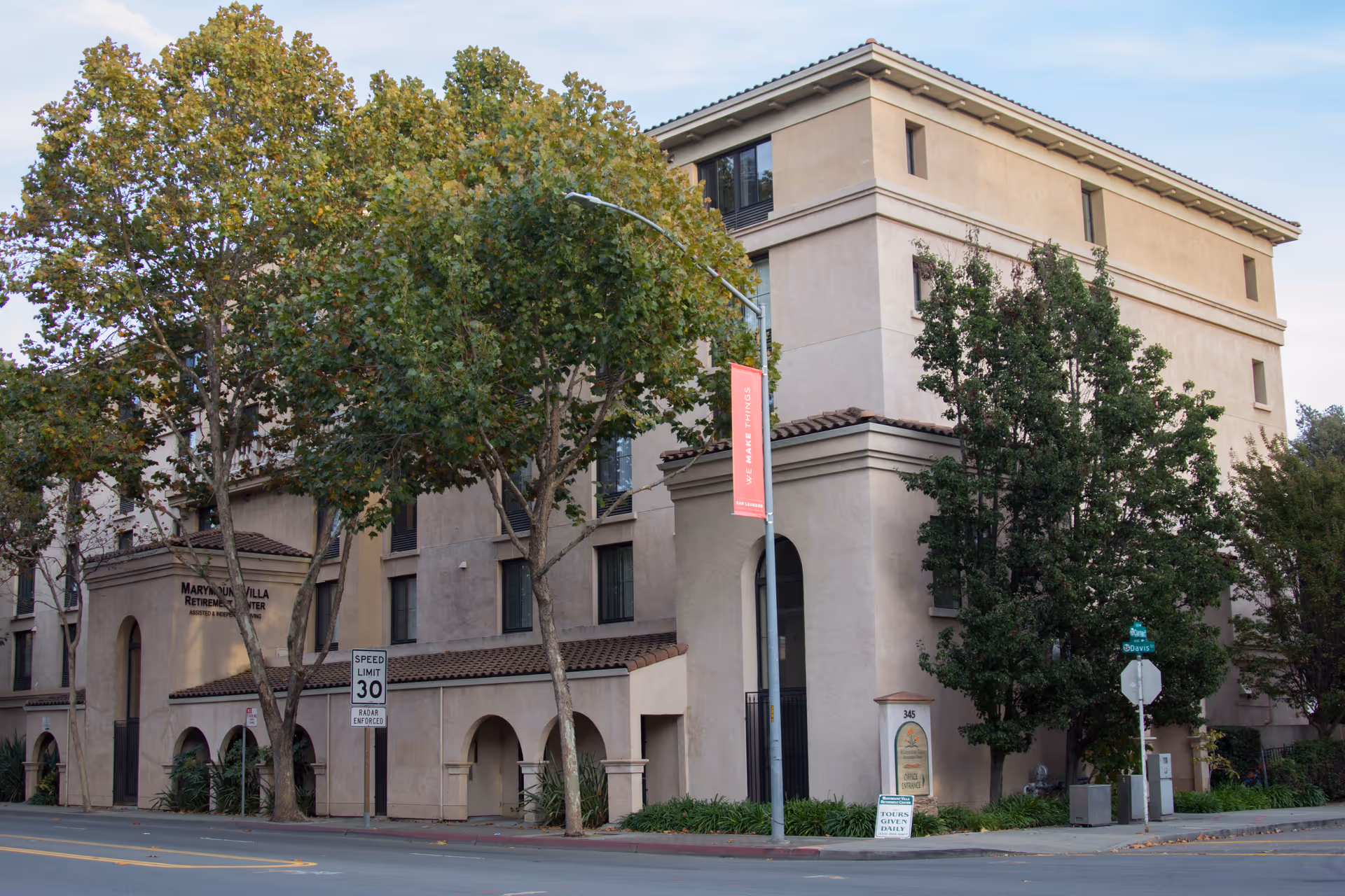Front exterior of the Marymount Villa senior living building, a multi-story beige stucco structure with trees and a street in front.