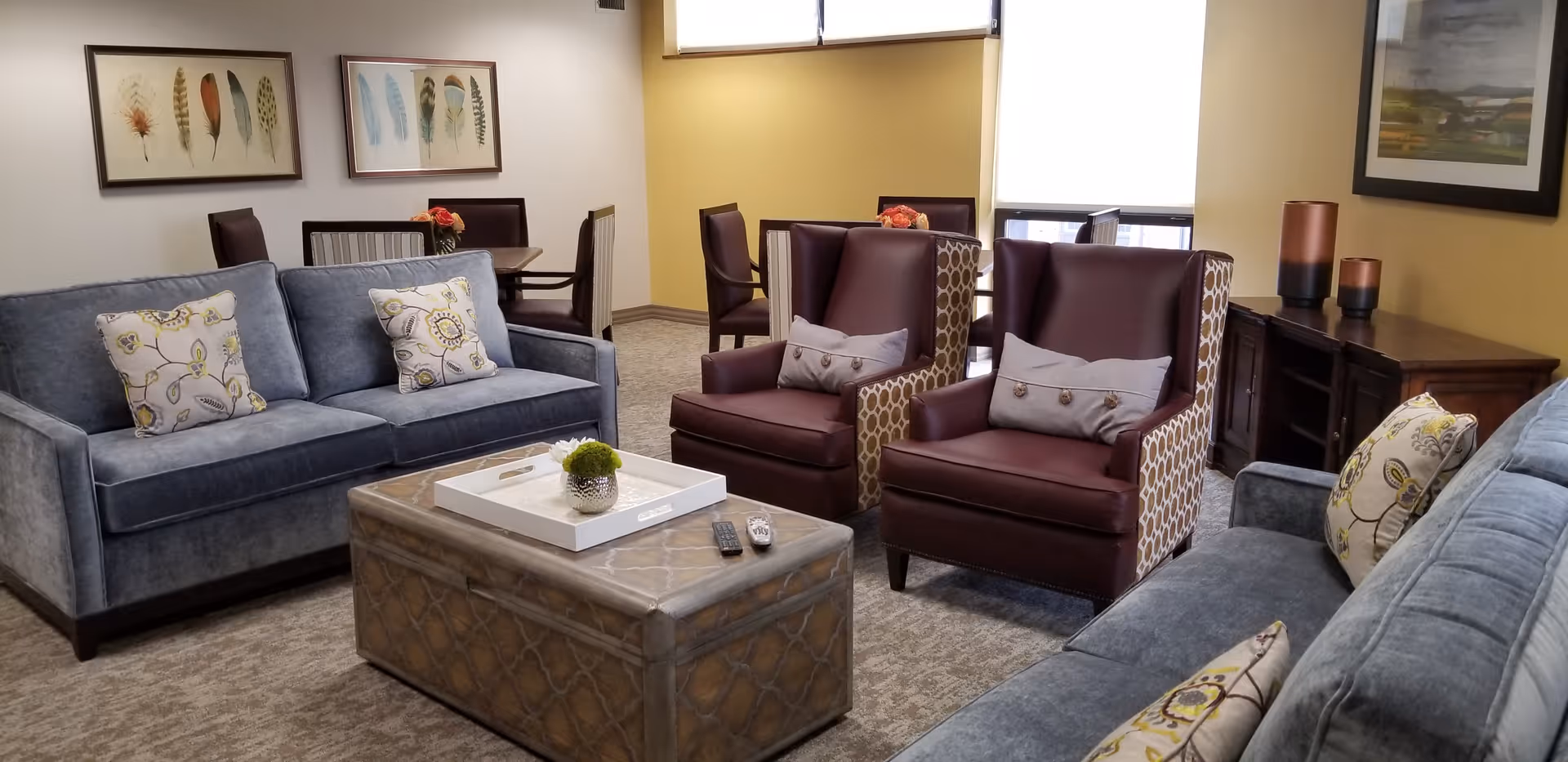 A cozy living room with two blue sofas and two maroon armchairs with patterned sides, arranged around a rectangular coffee table. The coffee table has a white tray with a small plant and two remote controls on it. The room has beige carpet, yellow and white walls, framed artwork on the walls, and a wooden sideboard with decorative items.