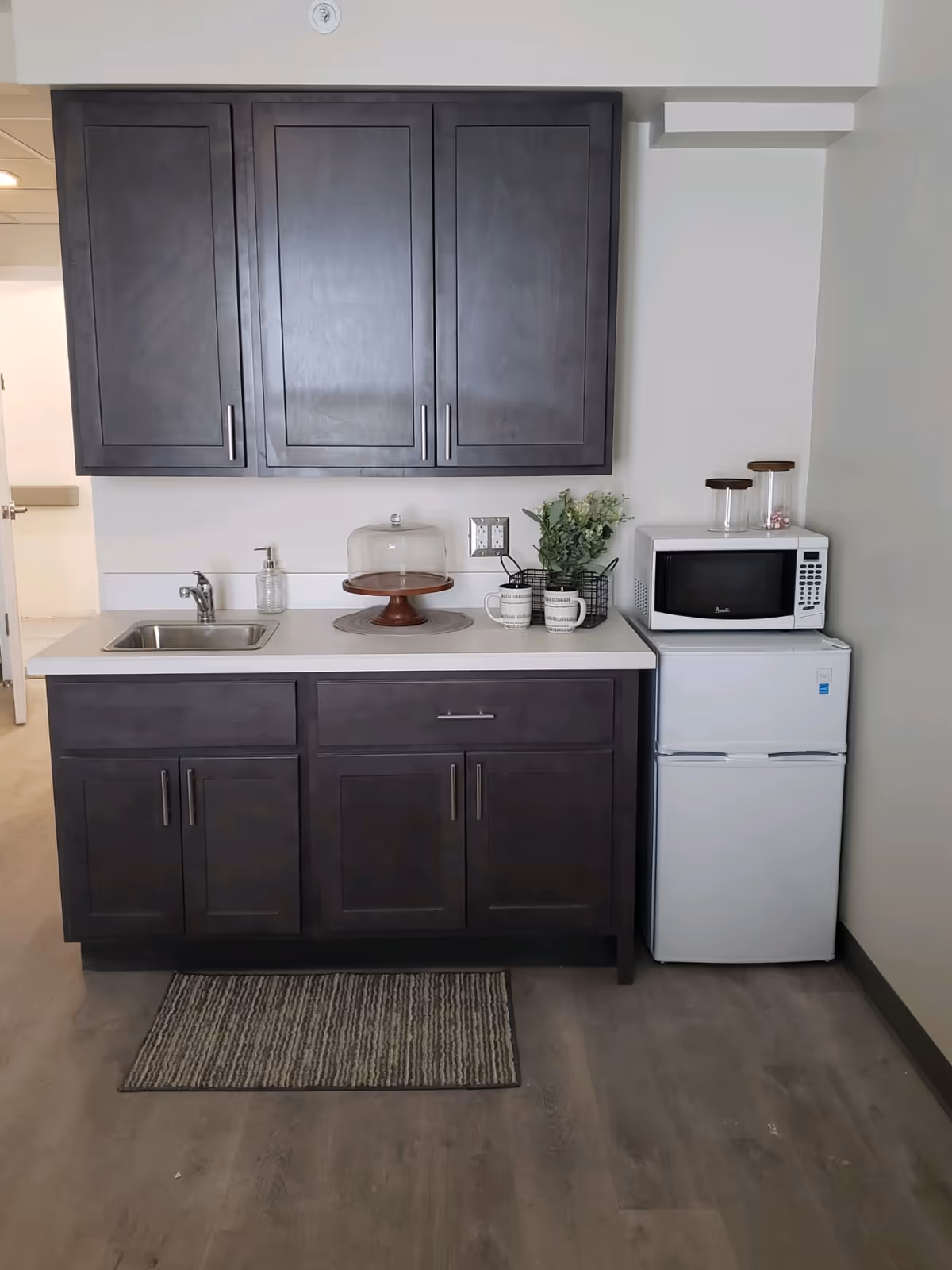 A small kitchenette area with dark wooden cabinets, a white countertop, a stainless steel sink, a soap dispenser, a cake stand with a glass cover, two mugs, a small plant in a wire basket, a white microwave, and a compact white refrigerator. A striped rug is placed on the wooden floor in front of the kitchenette.