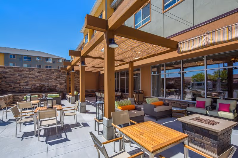 Outdoor patio area at Treeo Orem with wooden tables and chairs, cushioned lounge seating with colorful pillows, a fire pit, and a pergola providing shade. The area is surrounded by a stone wall and the building's exterior with large windows is visible.