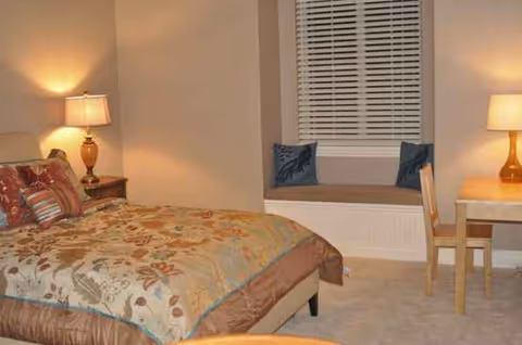 A furnished bedroom in a senior living facility with bed, window seat, and desk.