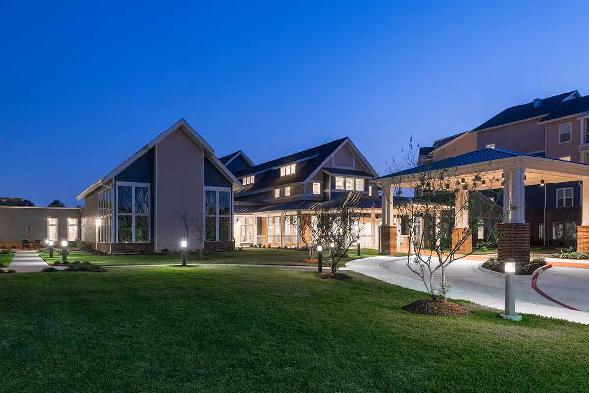 Exterior view of a retirement community building at dusk with a lit entrance, covered porte-cochère and landscaped lawn.